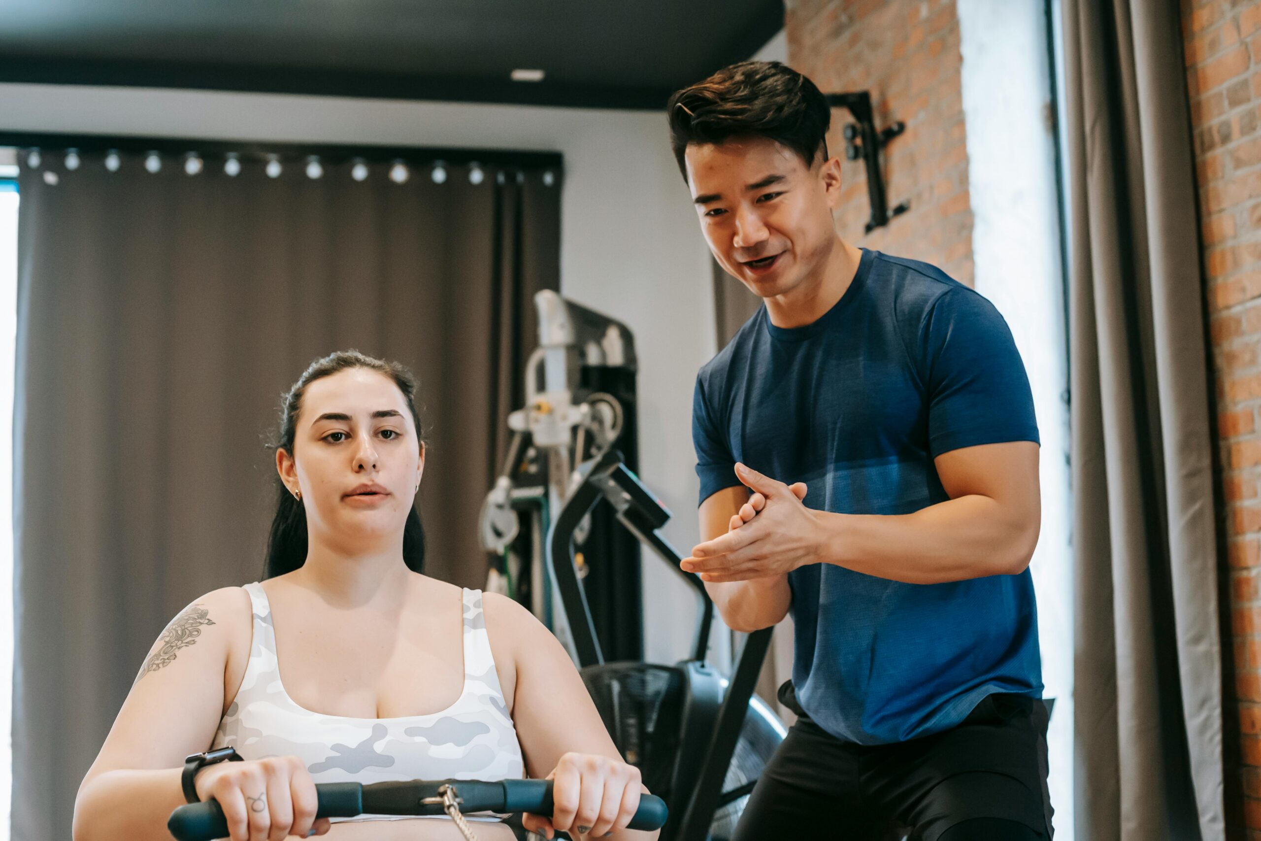 Low angle of positive Asian male trainer supporting plus size woman training on rowing machine during weight loss workout